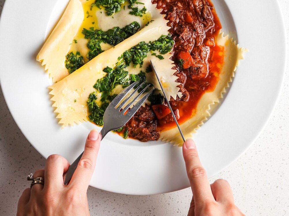Two hands are entering the bottom of the photo, one holding a knife the other a fork. They are cutting into a plate of pasta. On the left are wide, sheets of pasta topped with a green gremolata sauce. On the right, is a red beef ragu sauce.