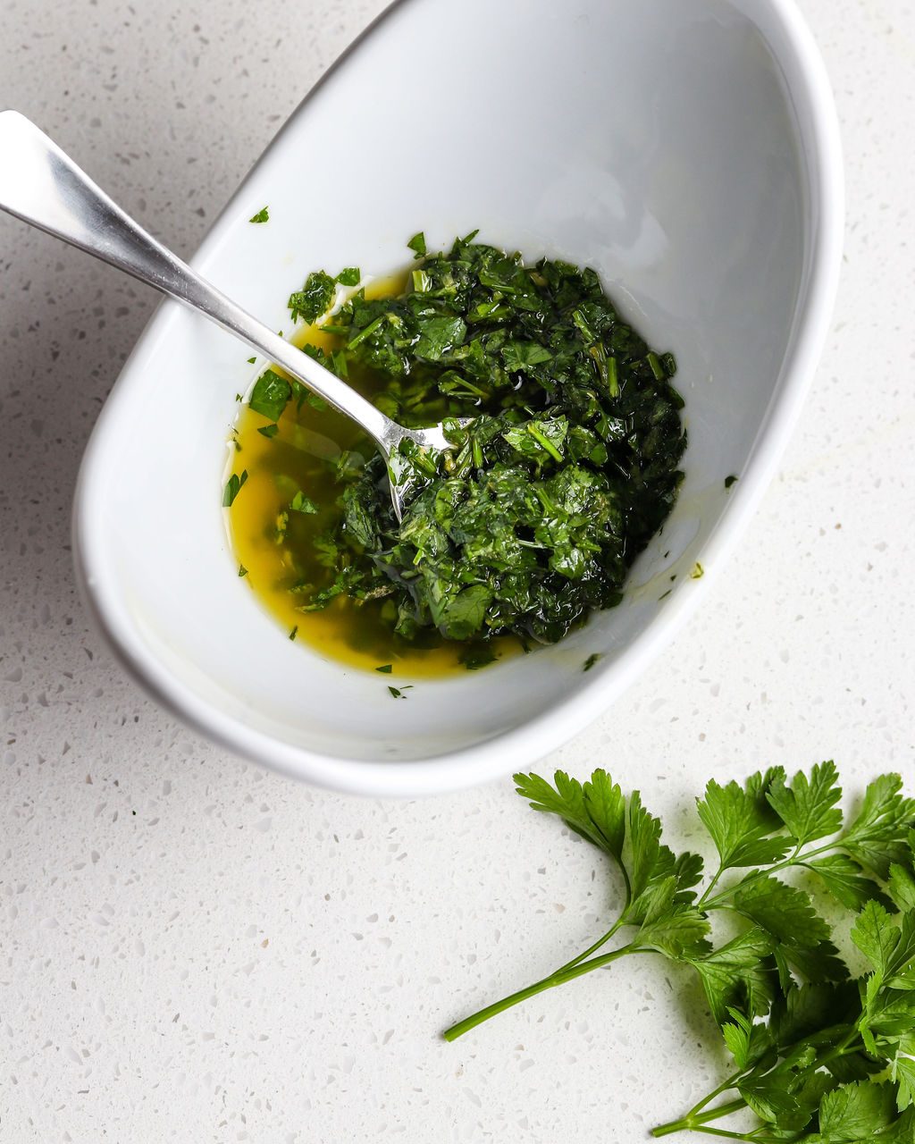 A white bowl holds a green gremolata sauce with chopped parsley, lemon juice, and salt and pepper. The bowl is sitting on a white surface. In the bottom righthand corner are some green parsley leaves.