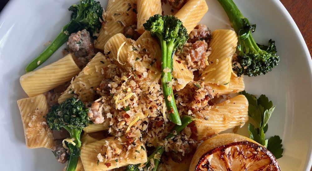 A shallow white bowl holds a mound of homemade rigatoni pasta, cooked broccolini florets, bits of sausage, and toasted breadcrumbs. In the bottom right corner of the bowl, sits a lemon half, with caramelized edges.