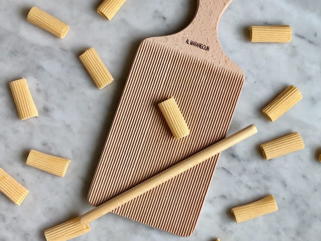 There is a light-colored wooden gnocchi board, with ridges and a handle at the top. Below the handle is a stamped name, "Al Marangoun." On top of the board. lays a wooden dowel and a piece of homemade rigatoni. The board sits on a white marble surface and there are other rigatoni pieces around the board.