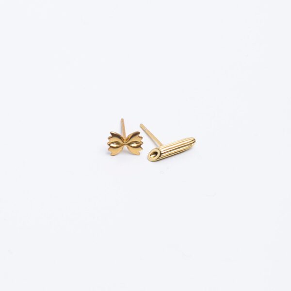 A pair of mismatched gold pasta earrings sits in the center upon a plain, white surface. We can see one farfalle (bowtie) earring and one ridged penne earring.
