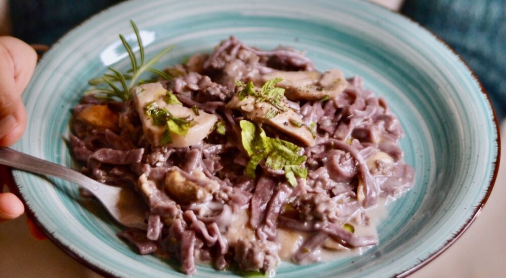 A teal ceramic plate holds a mound of reddish-purple pasta coated in a creamy mushroom sauce and topped with fresh herbs. In the background, we can see a hand holding the plate.
