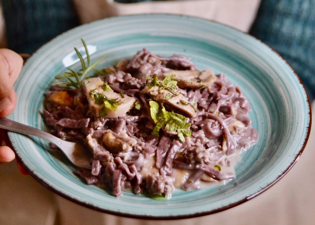 A teal ceramic plate holds a mound of reddish-purple pasta coated in a creamy mushroom sauce and topped with fresh herbs. In the background, we can see a hand holding the plate.