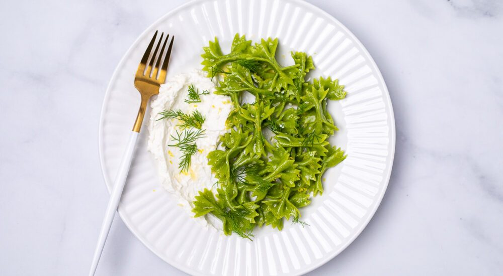 Spinach & dill farfalline pasta on a white plate. On the side we see some whipped goat cheese topped with green dill. There is a gold and white fork resting on the side of the plate.