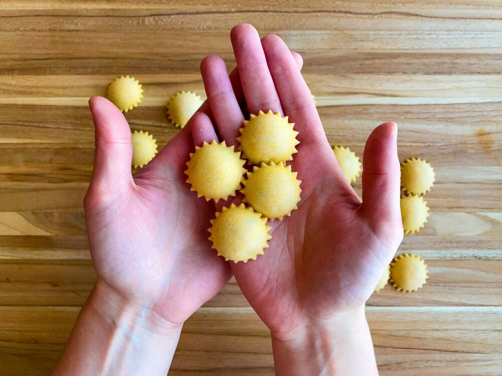 A pair of hands holds four anolini pastas; below we see a wooden surface with other anolini resting on top.