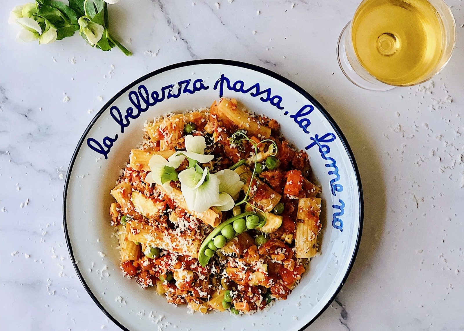 There is a bowl of rigatoni pasta with a tomato vegetable sauce, topped with parmesan shavings and pea tendrils. The bowl is white with a blue handpainted phrase that reads, "la bellezza passa, la fame no," which is Italian for, "beauty fade, hunger doesn't." In the top right corner we see a glass of golden white wine.