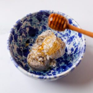 Blue splatterware bowl with three scoops of speckled gelato inside. A hand holds a wooden honey dipper, drizzling honey on top.