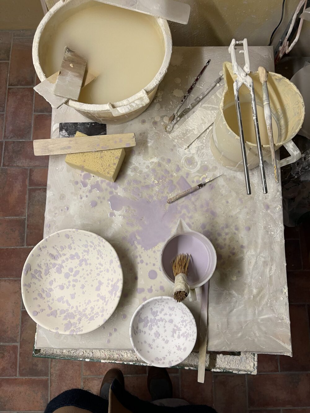 Splatterware in the making: there are two bowls painted with a light purple splatter. We see paint brushes and buckets full of paint.