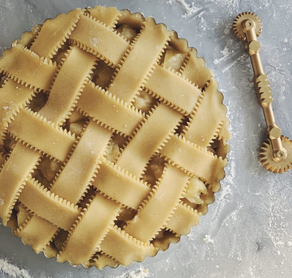 We see a latticed pie from above, with a warm tan color. The surface that it's sitting on has flour sprinkled over it and there is a brass pastry cutter with one wheel on each end.