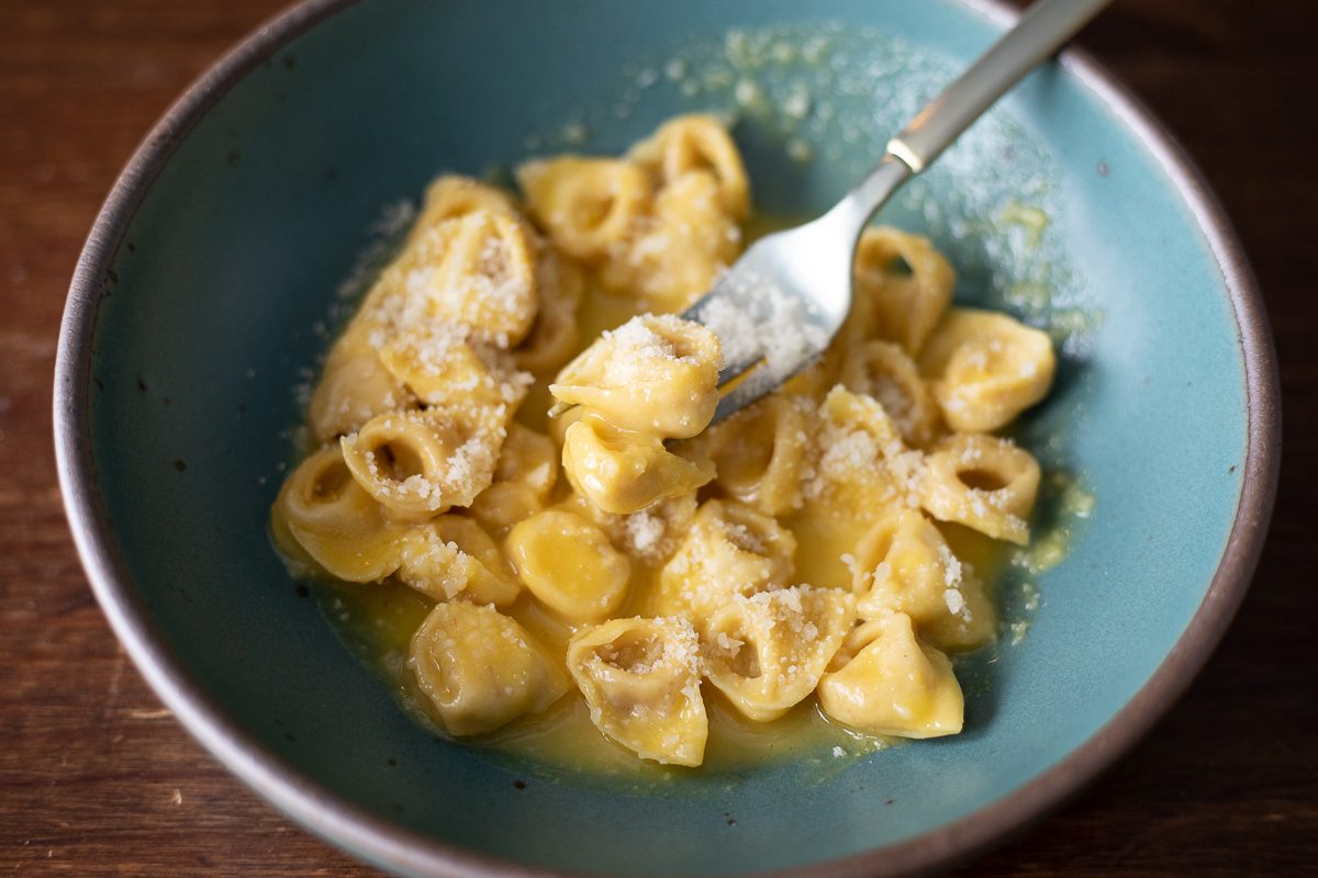 A teal low-bowl holds a heaping of golden tortellini with a creamy yellow sauce. We see a fork picking up a single tortellino. There is grated parmesan cheese on top.