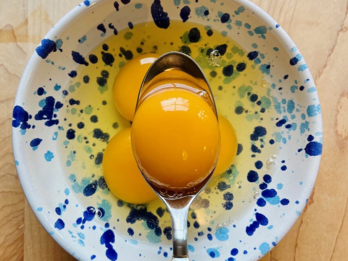 There is a blue splatterware bowl on a wooden surface holding three cracked eggs. Above the bowl we see a metal spoon holding a single orange egg yolk.