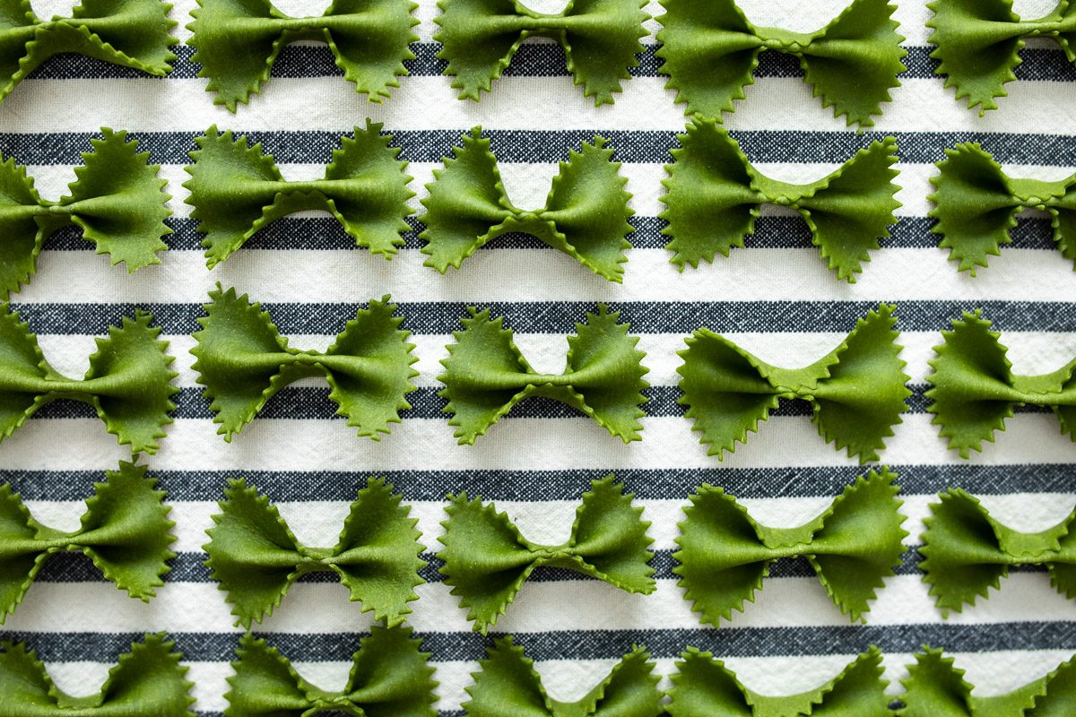 Green pasta bowties are lined up in neat rows. We see a blue striped tea towel underneath the pasta.